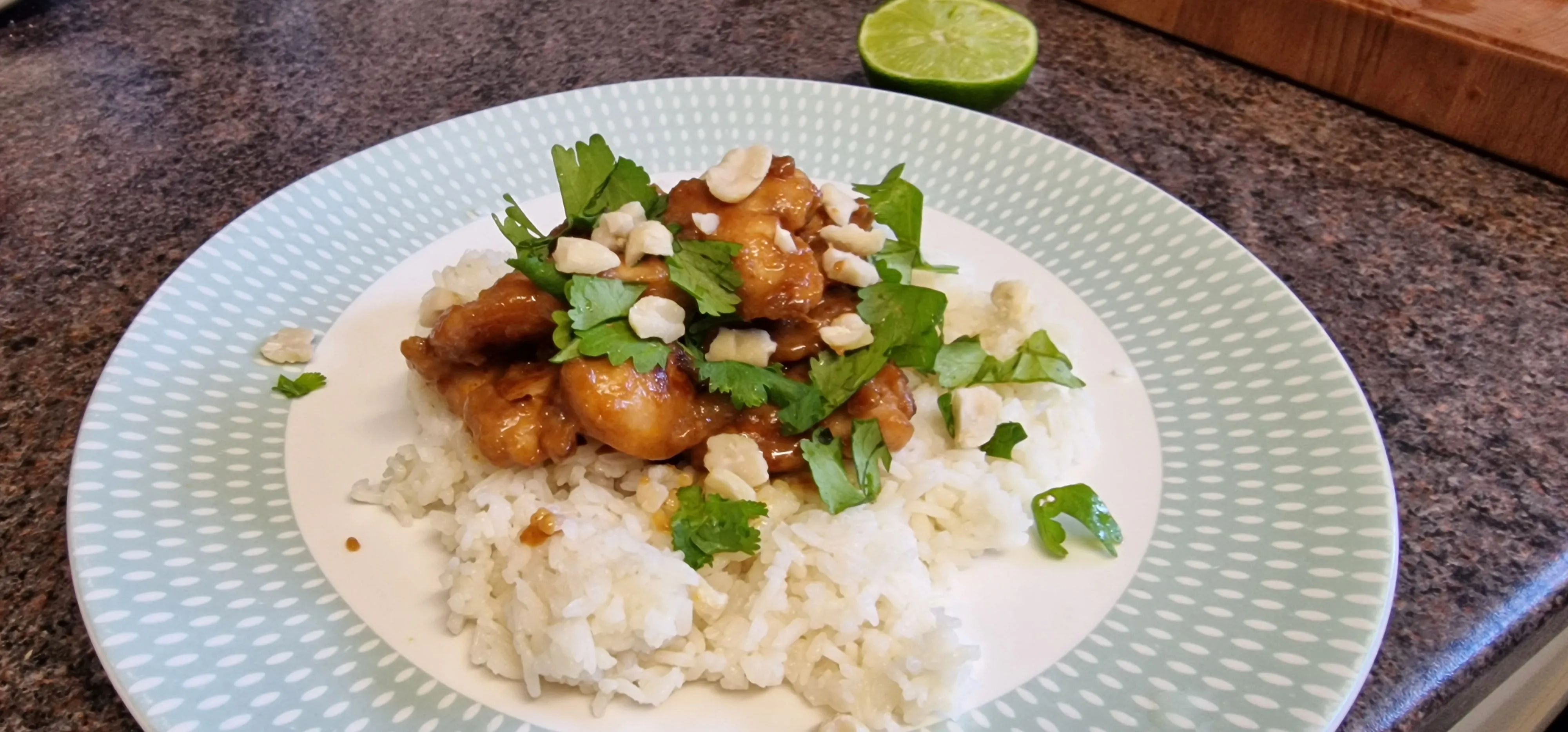 A plate of rice topped with glossy chicken, coriander, chopped peanuts and a lime half