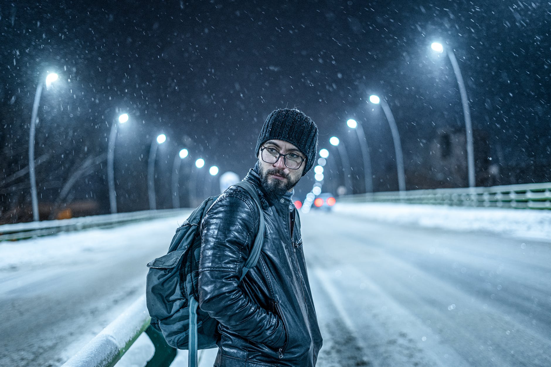a man in leather jacket carrying backpack while standing on the road during snow fall